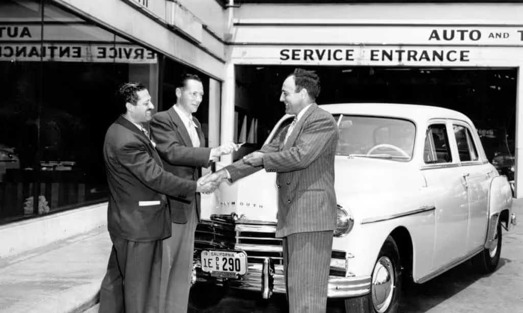 Black-and-white 1950s photo of salesmen shaking hands over a new Plymouth at a dealership service entrance